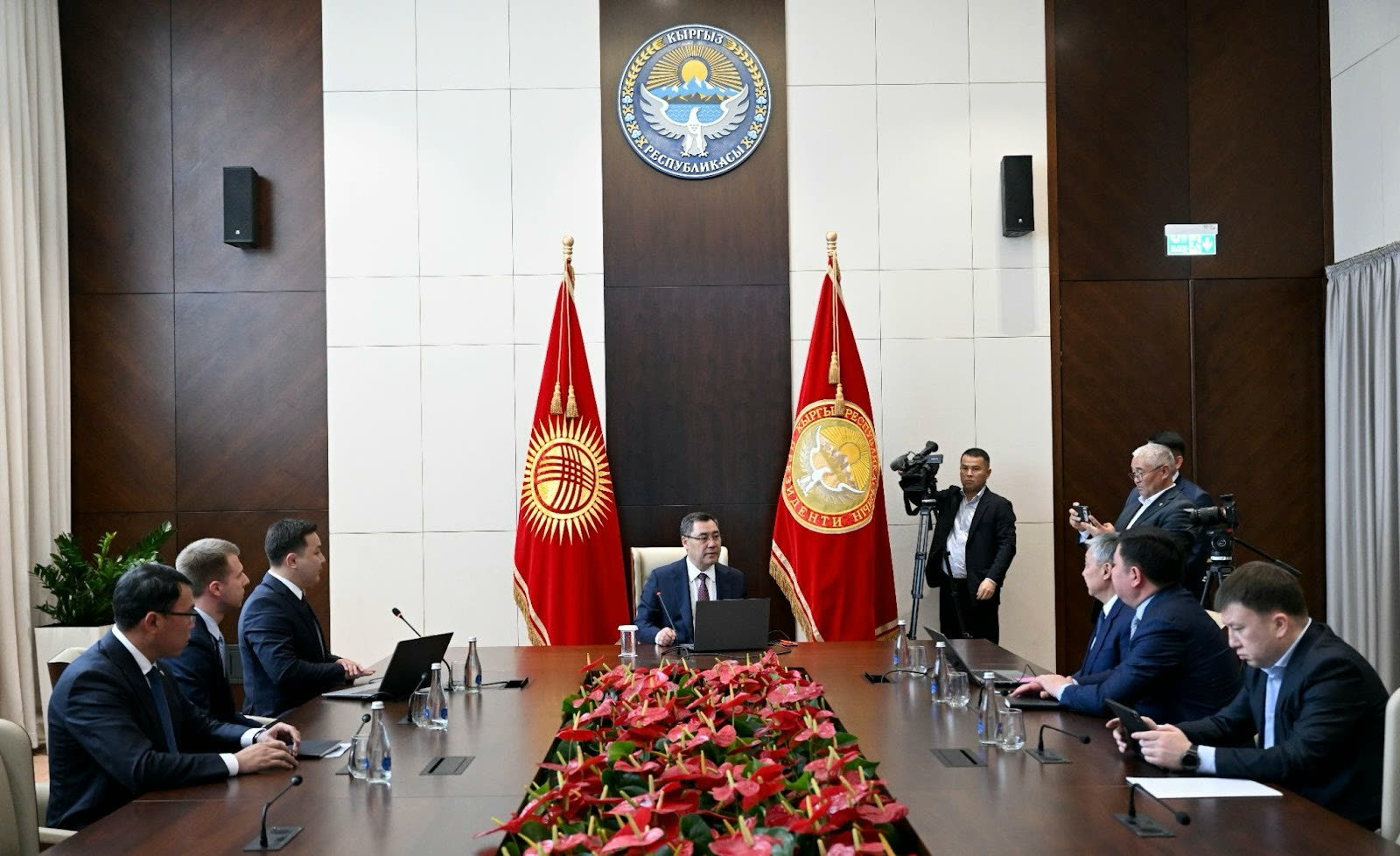 An official meeting room of the Kyrgyz Republic: delegates at a long table beneath the state emblem and national flags.